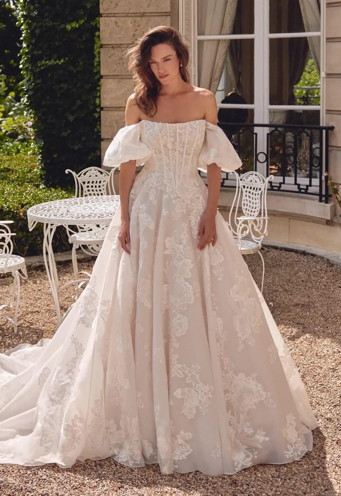 Model wearing an ivory floral lace wedding gown with a structured basque-waist corset, off-the-shoulder puff sleeves, and a full ball gown skirt.