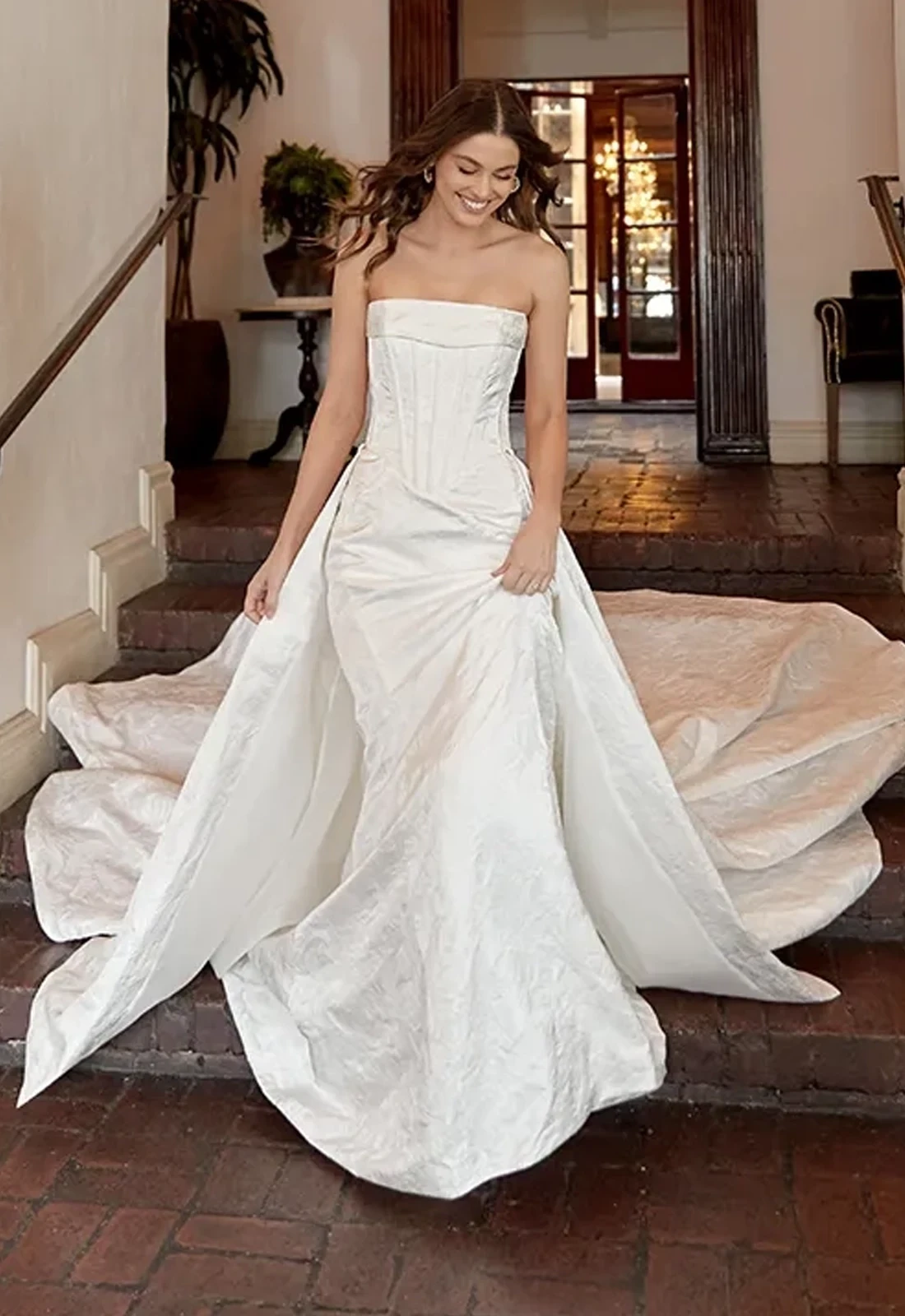 Model wearing a strapless ivory wedding gown with a structured corset bodice, fitted silhouette, and dramatic detachable overskirt with a long train.