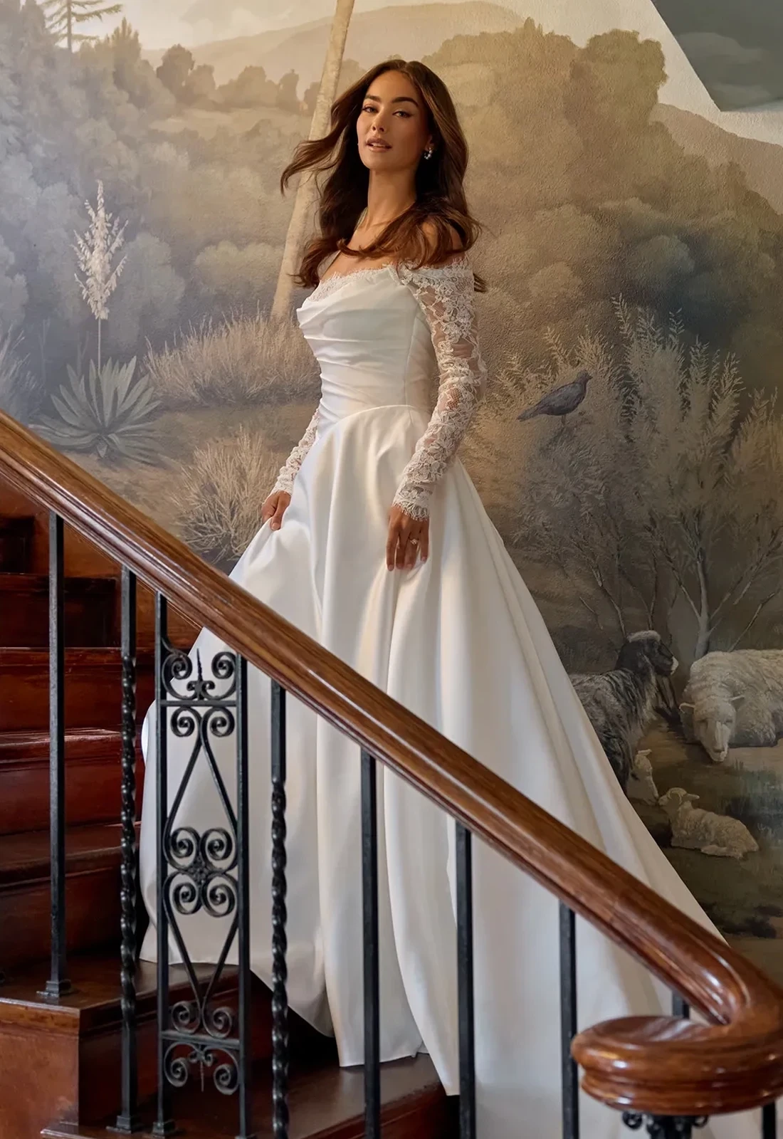 Model wearing an ivory satin wedding gown with long lace sleeves, an off-the-shoulder neckline, and a full flowing skirt.