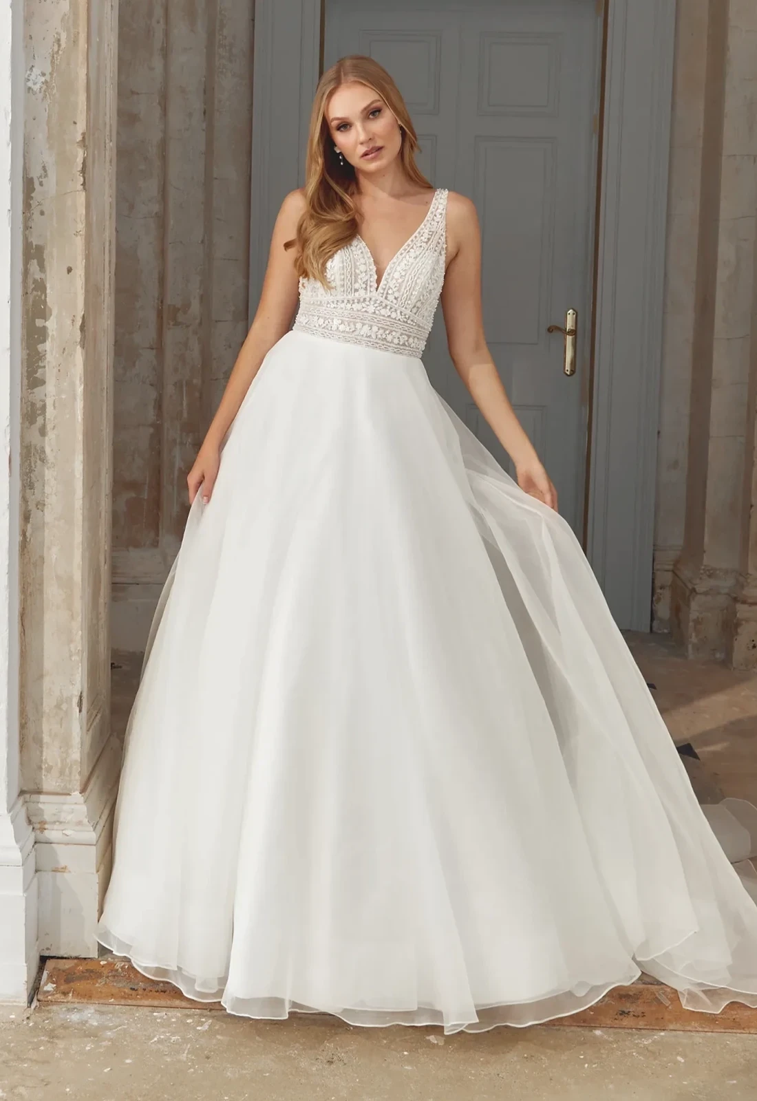 Model wearing an ivory A-line wedding gown with a deep V-neckline, intricately beaded lace bodice, and a soft flowing skirt.